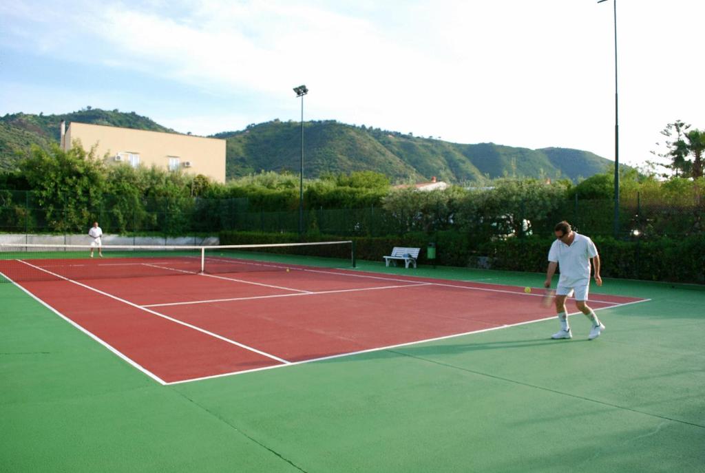 Tennisa laukums ar diviem spēlētājiem, kas nodarbojas ar spēli. Fons veido kalni un zāle, radot mierīgu atmosfēru.