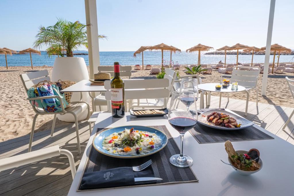 Pludmales restorāna galdiņš ar gourmet ēdieniem, sarkano vīnu un skaistu skatu uz jūru. Apvērstas sauļošanās vietas un palmas papildina relaksējošo atmosfēru.