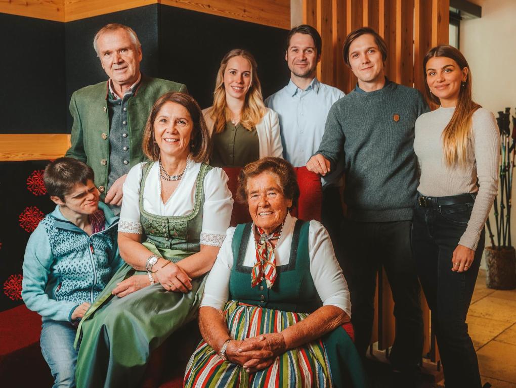 Grupas fotoattēls ar cilvēkiem tradicionālās tērpos, kas pozē pie sarkanā dīvāna. Attēls atspoguļo ģimenes saikni un kultūras mantojumu.