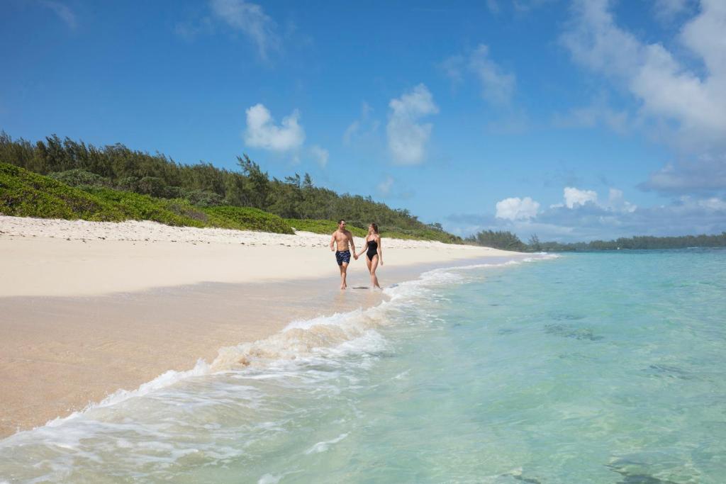 Pāris pastaigā gar skaistu pludmali, kur zils ūdens maigi viļņojas pie kājām. Fons ir zaļi krūmi un zils debesis ar dažām mākoņainām vietām.