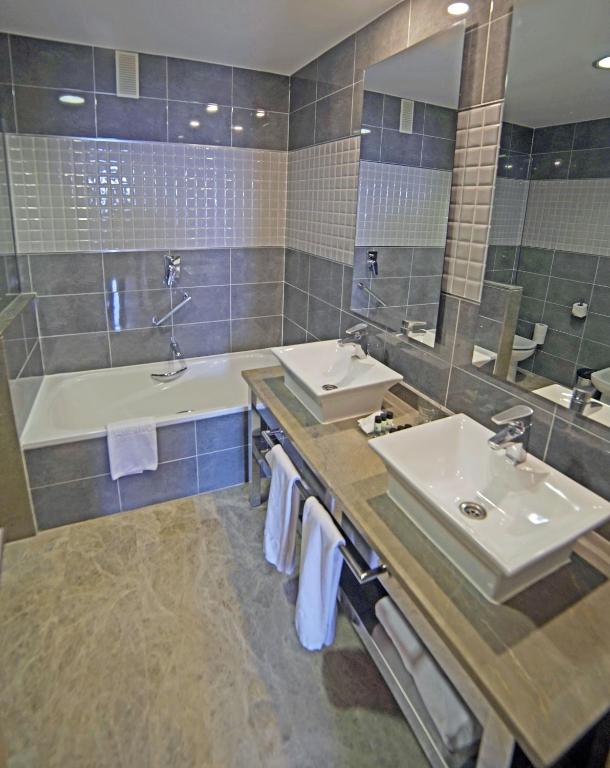 Mūsdienīgs vannas istabas interjers ar vannu, divām izlietnēm un spoguļiem, veidots pelēkās un baltās krāsās. Eleganta un funkcionāla telpa ar modernām akcesuāriem.