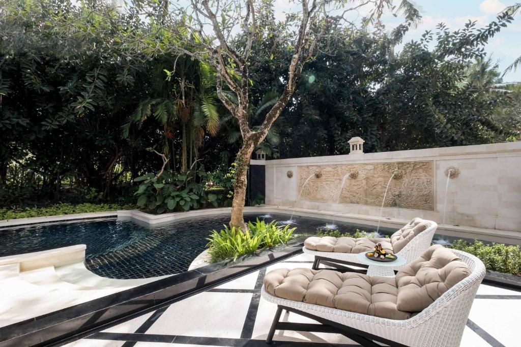 Relaksējošs dārza stūris ar baseinu, greznām atpūtas krēslām un dekoratīvu strūklaku, ieskauts zaļo augu ainavā. Ideāla vieta mierīgām brīvā laika stundām.