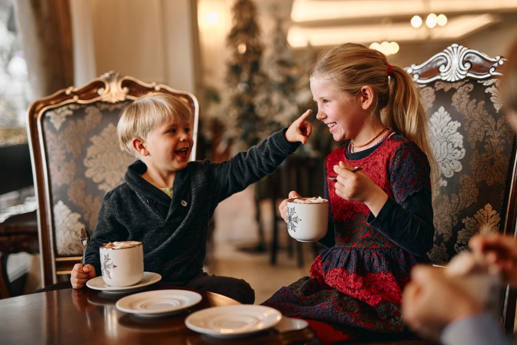 Bērni sēž kafejnīcā, izbaudot kakao ar putukrējumu. Zēns ar prieku norāda uz meiteni, kura smaida un turot krūzi rokā.
