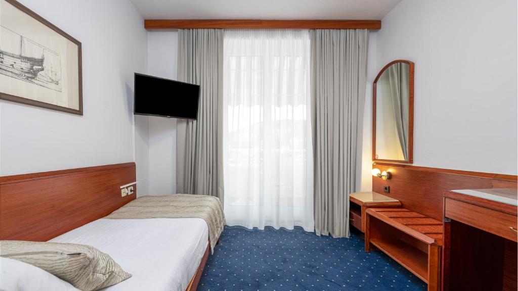 Mūsdienīga viesnīcas numura iekārtojums ar televizoru, gultu un lieliem logiem, kas nodrošina dabisko apgaismojumu. Numurā ir arī spogulis un koka mēbeles.