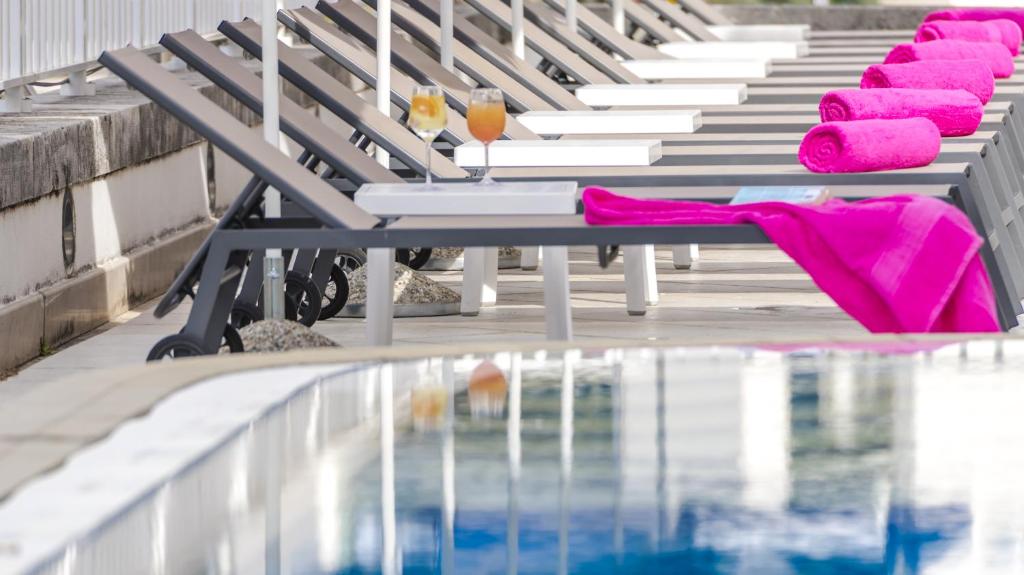 Baseina zona ar saules gultām, uz kurām novietoti rozā dvieļi, un galdiņiem ar dzērieniem. Ūdens spīd saulē, radot relaksējošu atmosfēru.