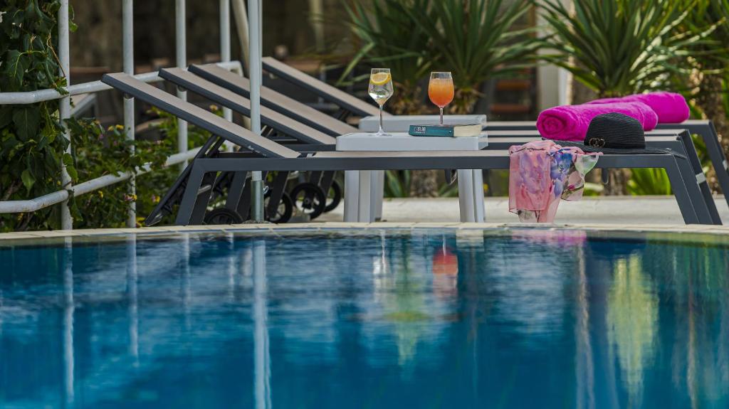 Baseina zona ar sauļošanās krēsliem, glāzēm ar dzērieniem un rozā dvieļiem, radot relaksējošu atmosfēru. Ūdens atspīd, piedāvājot mierīgu vidi atpūtai.