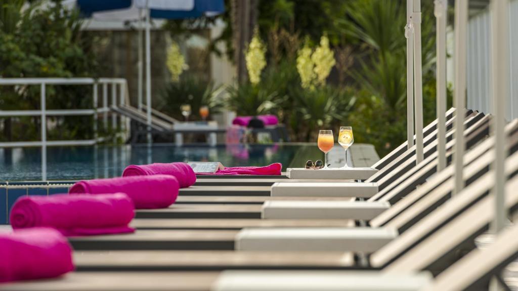 Atpūtas zona pie baseina ar rozā dvieļiem un divām dzērienu glāzēm uz galda. Svaiga zaļums un saulains laiks rada relaksējošu atmosfēru.