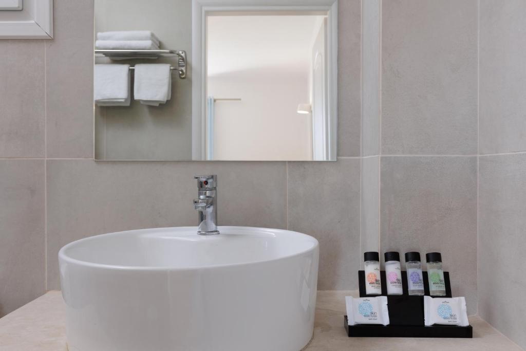 Mūsdienīgs vannas istabas izskats ar baltu izlietni un higiēnas piederumiem, kas sakārtoti melnā statīvā. Spogulī redzams papildu telpas elements.