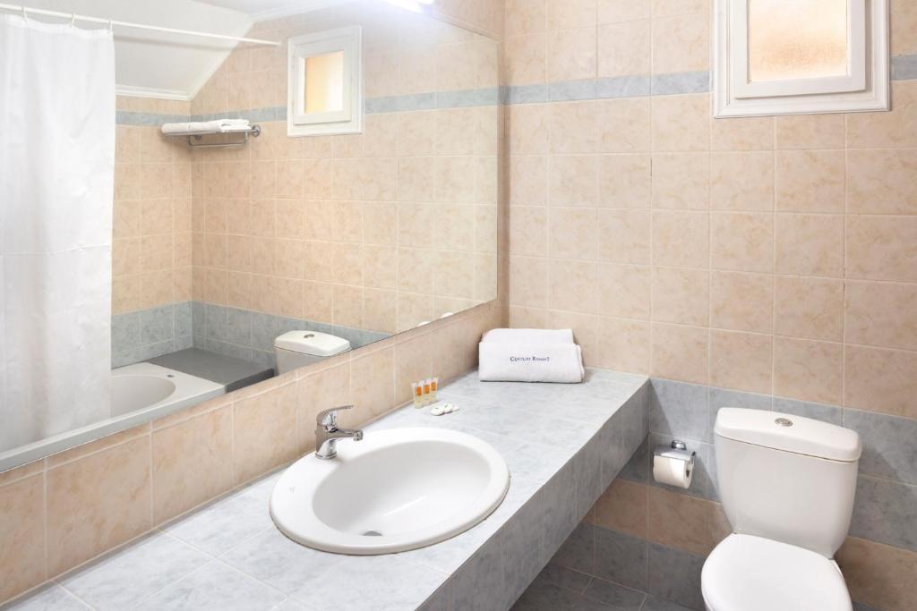 Mūsdienīga vannas istaba ar gaišām flīzēm, plašu spoguli un vannu. Tajā ir arī tualete un neliela darba virsma ar izlietni.