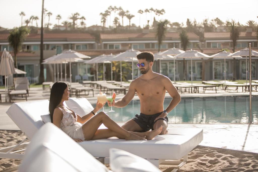 Pāris bauda dzērienus pie baseina, atpūšoties saulainā pludmales vidē. Ideāls brīvdienu mirklis, kas ietver relaksāciju un romantiku.