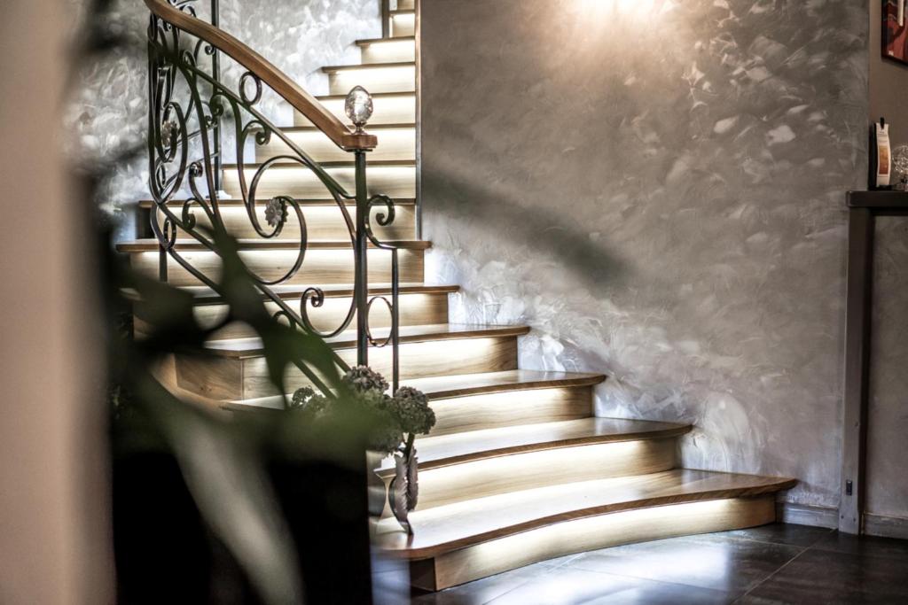 Eleganta kāpnes ar koka pakāpieniem un metāla margām, izgaismotas ar siltu gaismu, radot mājīgu atmosfēru. Fons ir viegls un moderns, ar nelieliem dekoratīviem elementiem.