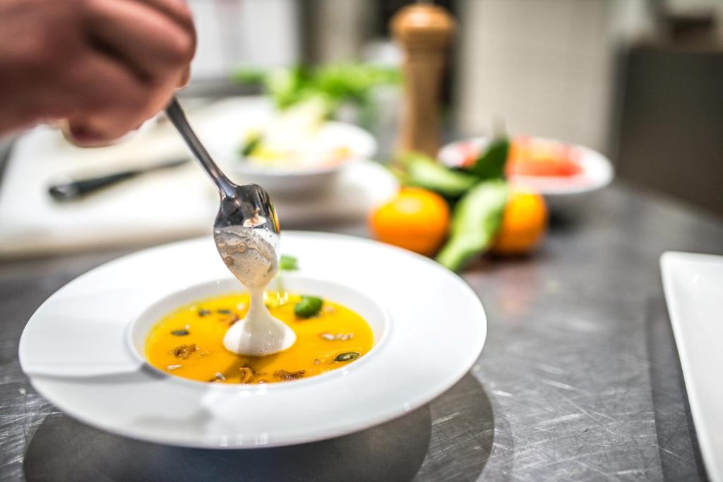 Siltā zupa ar krēmīgu piedevu tiek pasniegta baltā traukā, kam apkārt ir svaigi dārzeņi un garšaugi. Šī kulinārā aina izstaro siltumu un radošumu.