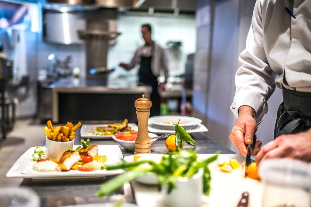 Šefpavārs sagatavo ēdienu profesionālā virtuvē, kur redzamas gatavošanas sastāvdaļas un pasniegšanas plāksnes. Aiz viņa redzams cits pavārs, kas strādā.