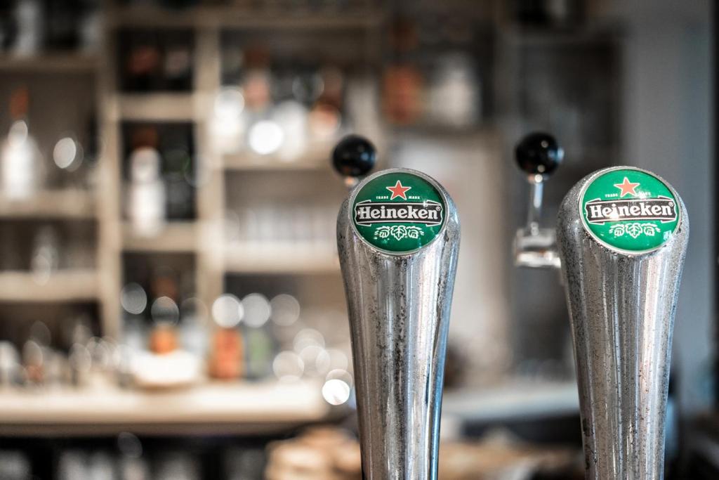 Divas Heineken alus krāni bārā, ar izplūdušu fona, kur redzami dažādi dzērieni.