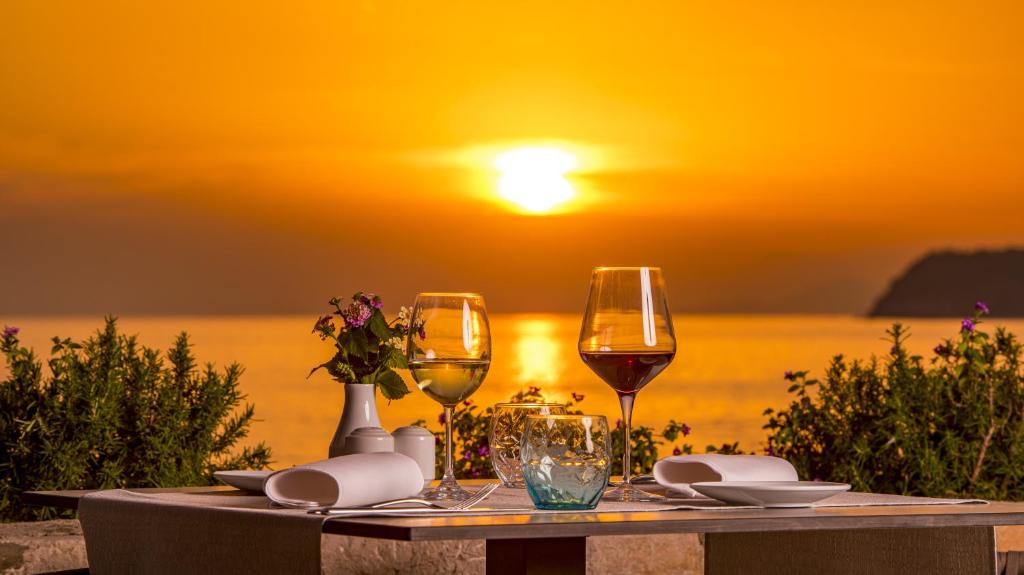 Romantiska vakariņas pie jūras ar saulrietu, kur uz galda ir vīna glāzes, ziedi un eleganti galda piederumi.