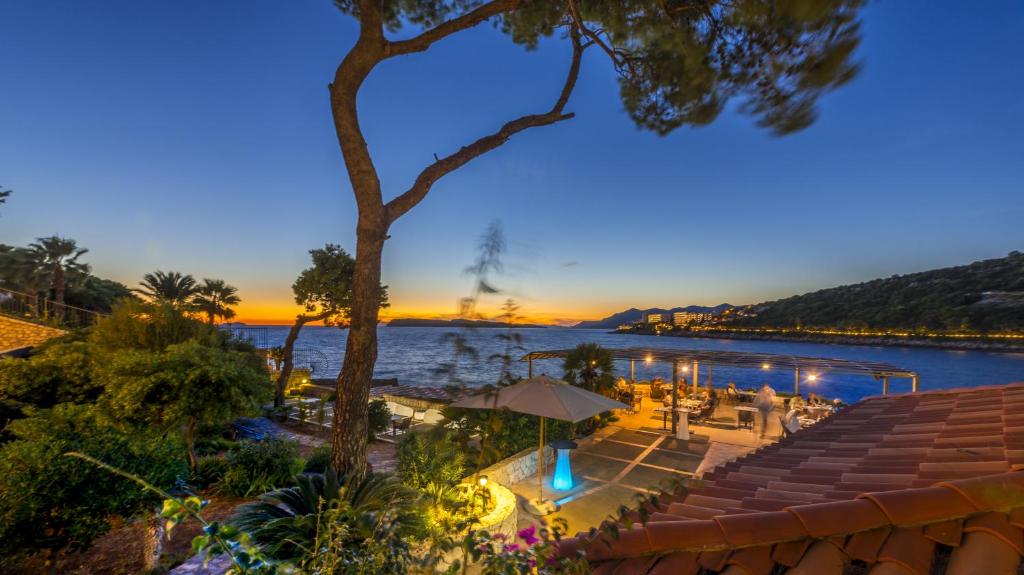 Saulrieta skats pie pludmales ar palmām un restorānu terasi, kas atrodas pie ūdens. Fons ir kalni un mierīgs jūras ūdens, radot relaksējošu atmosfēru.