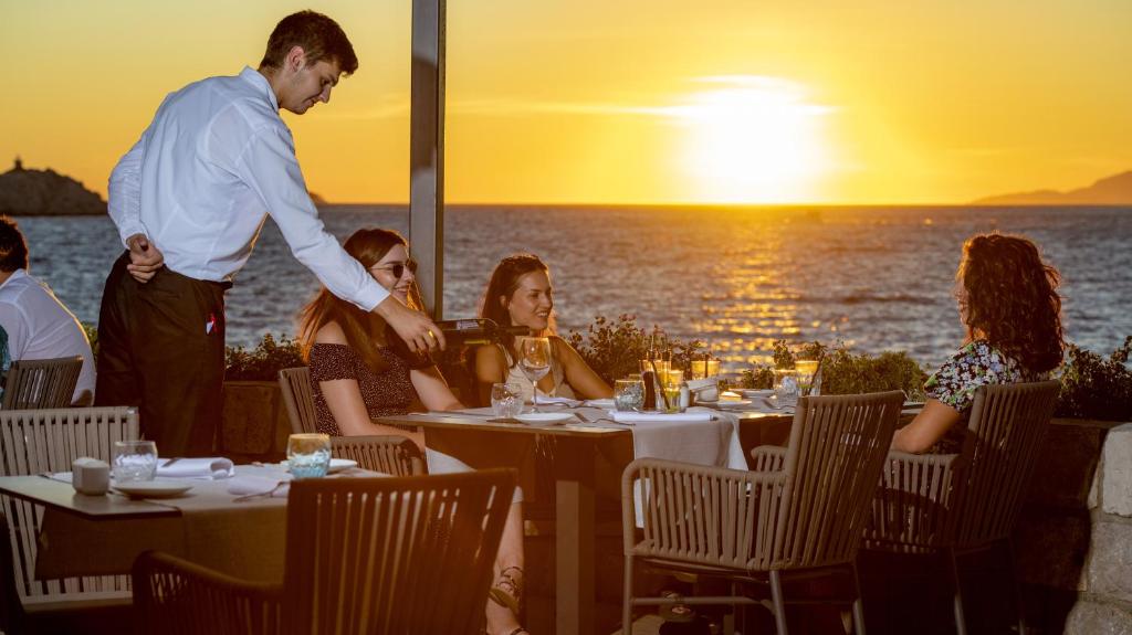 Saulriets pie pludmales restorāna, kur viesmīlis apkalpo trīs sievietes, kas bauda vakariņas ar skatu uz jūru. Atmosfēra ir relaksējoša un romantiska.