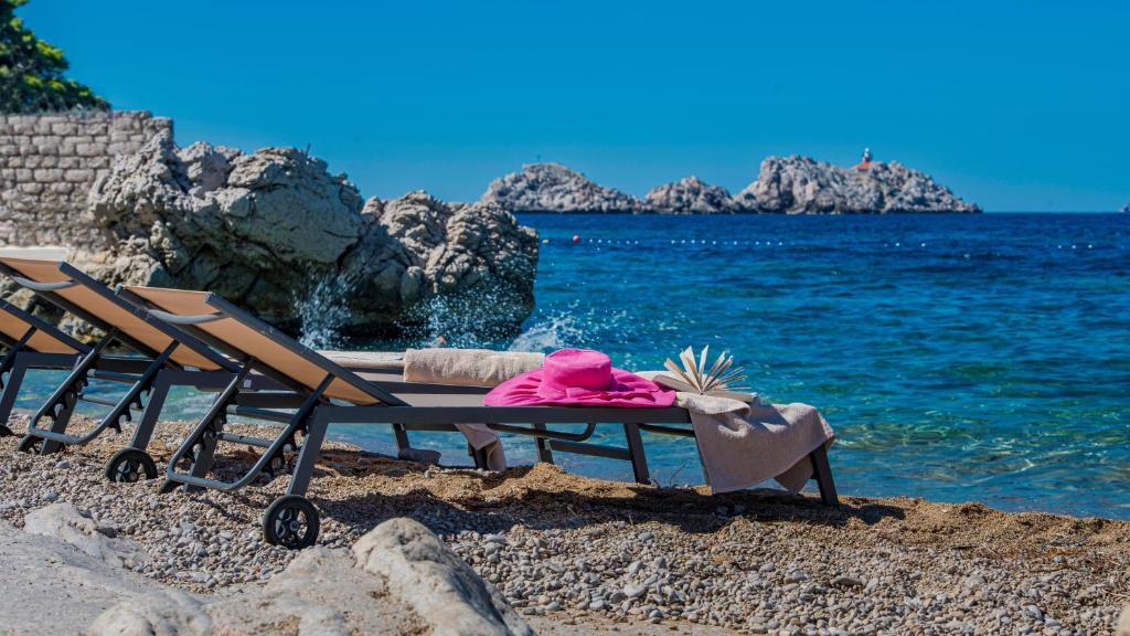 Atpūtas krēsli pie pludmales ar rozā cepuri un dvieli, fons ar skaidru zilo jūru un akmeņainiem krastiem. Ideāla vieta relaksācijai un atpūtai pie ūdens.