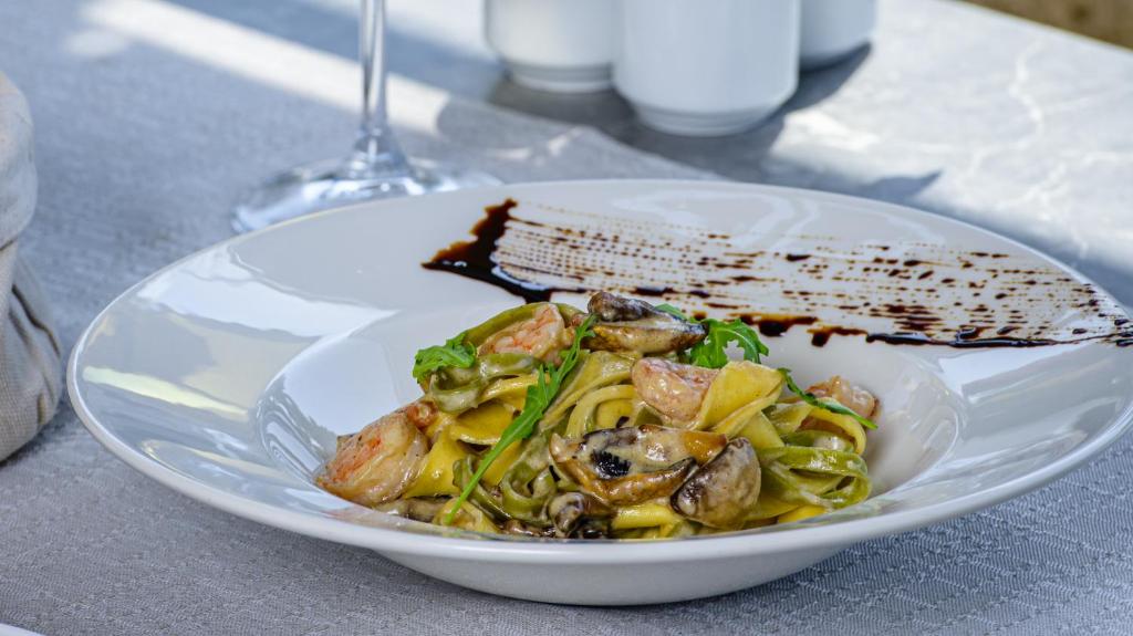 Pasta ar garnelēm un sēnēm, pasniegta uz elegantas baltas šķīvja ar dekoratīvu mērci. Fons ir neitrāls, ar glāzi vīna un citām ēdiena pasniegšanas detaļām.