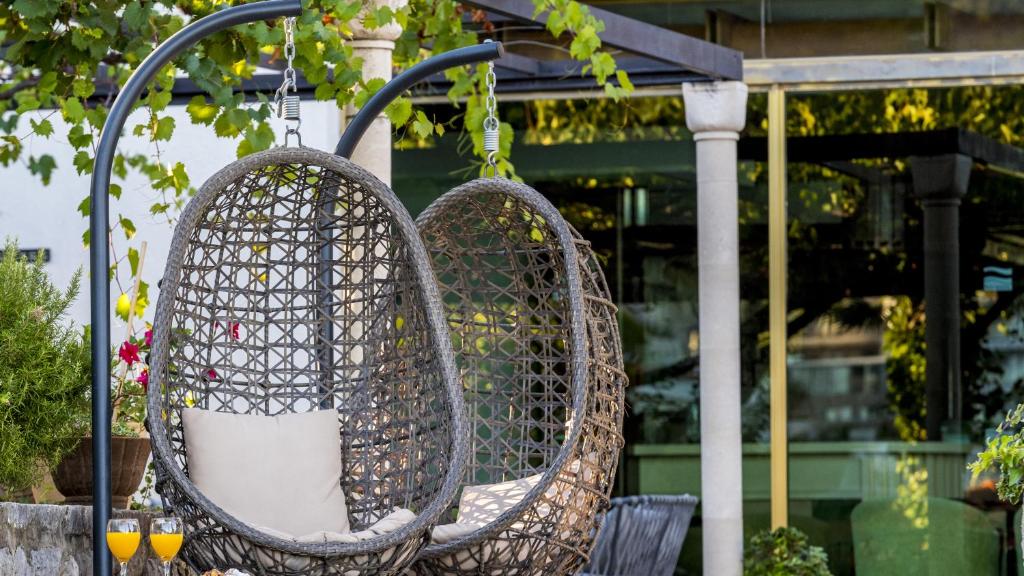 Šūpuļkrēslu komplekts ar mīkstiem spilveniem un dzērieniem, novietots dārzā starp augiem un stikla ēku. Ideāla vieta relaksācijai un atpūtai.