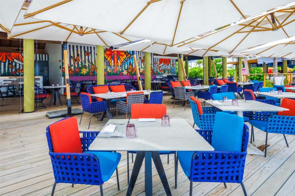 Krāsains restorāna terases interjers ar sēdvietām un saules jumtiem, izceļot tropisko atmosfēru un mākslinieciskos sienu gleznojumus.