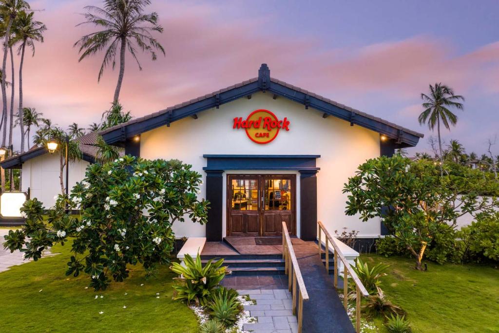 Hard Rock Cafe ieeja, apzaļumota vide ar palmām un ziediem, pie saulrieta. Kafejnīca ar modernu arhitektūru un izteiksmīgu zīmi.