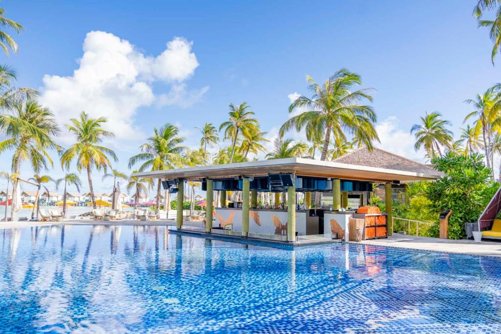 Tropu baseins ar palmām un bāru, kas atrodas pie ūdens, nodrošina relaksējošu atmosfēru. Sauļošanās krēsli un saulainas debesis papildina tropu noskaņu.