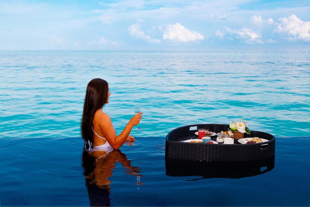 Sieviete, kas sēž ūdenī, tur glāzi dzēriena, kamēr blakus ir peldējoša galda ar ēdieniem un dzērieniem. Fons veido skaista zilā jūra un debesis.