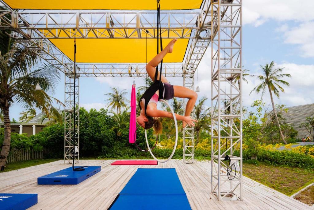 Gaisa akrobātika uz riņķa zem saules, ar dzeltenu nojumi un kokiem fonā. Vingrotāja izpilda izsmalcinātu kustību uz akrobātikas aprīkojuma.