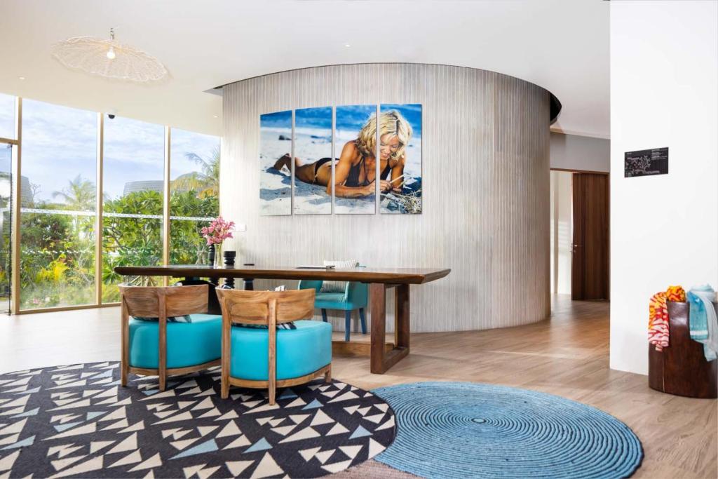 Mūsdienīgs dzīvojamās telpas interjers ar apaļu sienu, kurā atrodas gleznas, un elegantu mēbeļu kombināciju, kas ietver koka galdu un krēslus ar spilgtu zilo apdari. Telpa ir piepildīta ar dabīgu gaismu un rotāta ar ziediem un paklājiem.