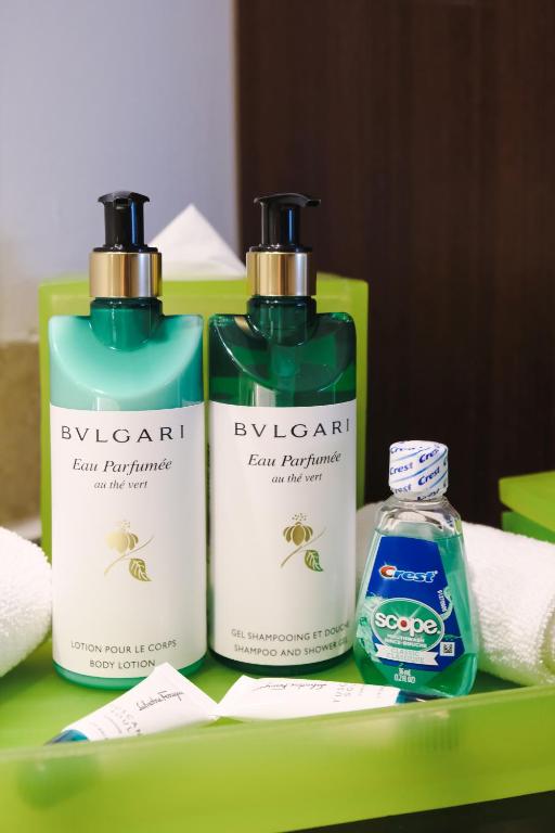 Vannas istabas aksesuāri, tostarp Bvlgari ķermeņa losjons un šampūns, kā arī Crest zobu skalošana, izvietoti uz zaļas virsmas ar dvieļiem.