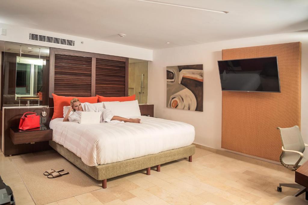 Mūsdienīgs viesnīcas numurs ar plašu gultu, kurā atpūšas sieviete ar klēpjdatoru. Interjerā ir siltas krāsas un moderns dizains, kas rada mājīgu atmosfēru.