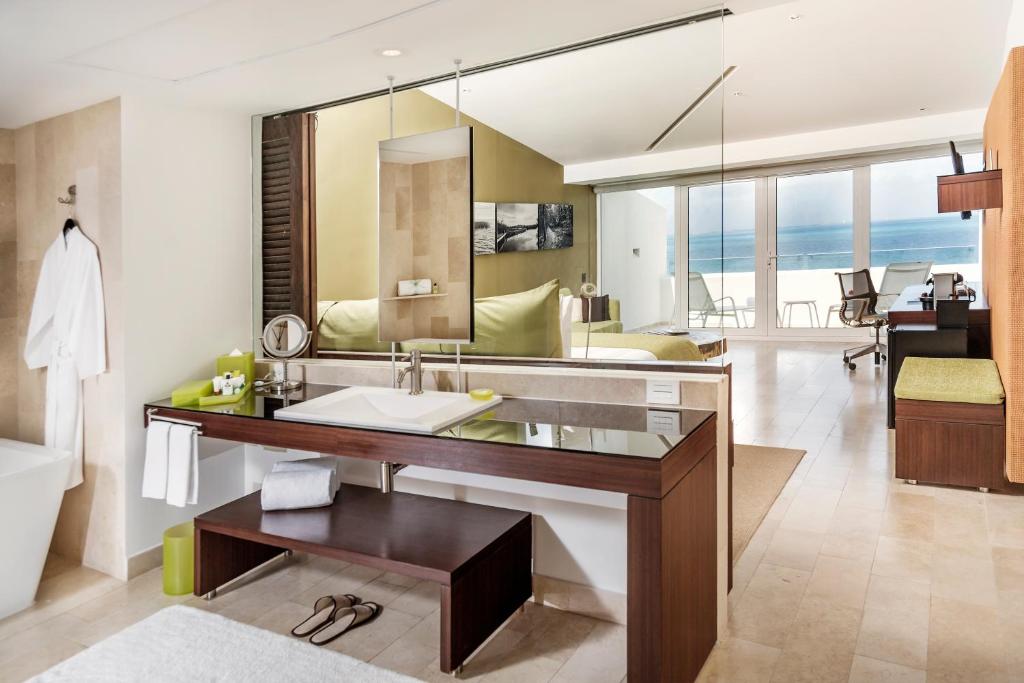 Moderns viesnīcas numurs ar vannas istabu, kurā ir eleganta izskata izlietne, spogulis un brīva gaisma. Skats uz jūru caur lieliem logiem, kas piešķir telpai plašuma sajūtu.