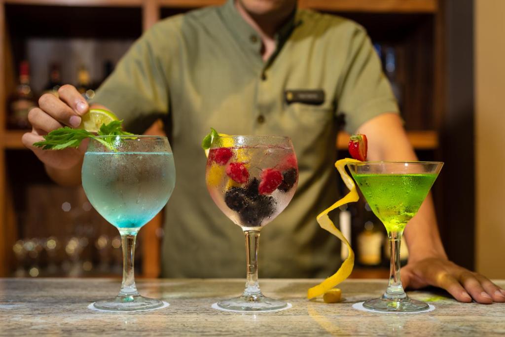 Dzerienu pagatavošana bārā, kur redzami trīs krāsaini kokteiļi - zilā, caurspīdīgā un zaļā krāsā, ar augļiem un garšaugiem dekorētiem.