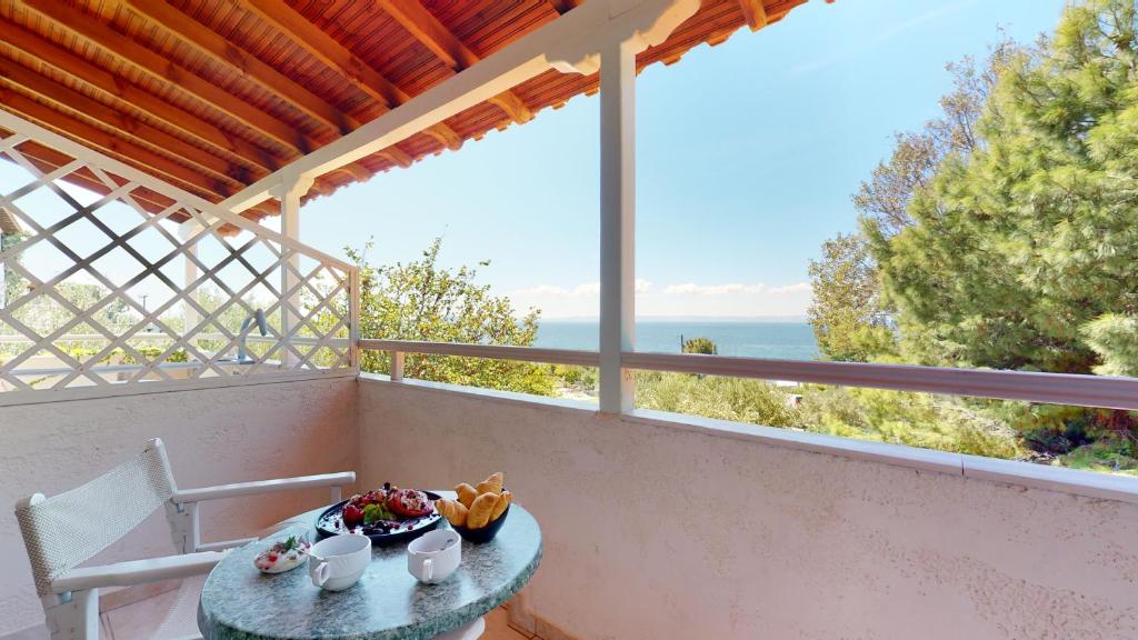 Balkons ar skatu uz jūru, uz kura atrodas galds ar brokastu klātbūtni, tostarp augļiem un maizi, un krēsls. Daba apkārt ir zaļa un saulaina.