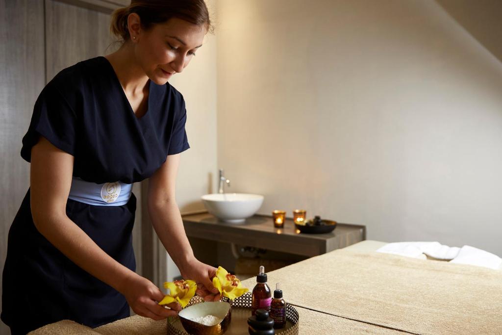 Skaistumkopšanas speciāliste sagatavo masāžas telpu, novietojot ziedus un eļļas uz masāžas galda. Telpā ir mierīga atmosfēra ar svecēm un elegantu interjeru.