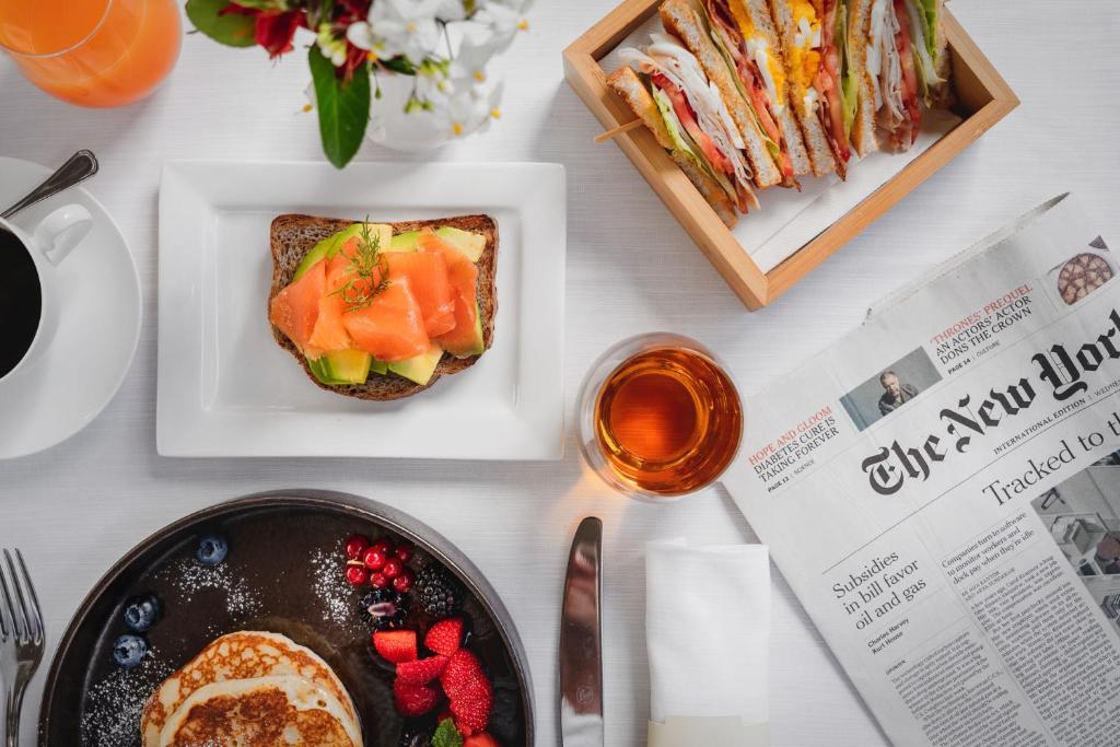 Svaiga brokastu plate, kurā ir pankūkas ar ogām, avokado un laša uz maizes, kā arī sviestmaizes un tase kafijas. Blakus atrodas laikraksts un glāze tējas.