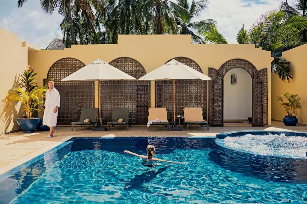 Luksusa spa ar baseinu, kurā peld bērns, un vīrietis baltā halātā stāv blakus, iepriecināts, apzaļumots ar palmām.