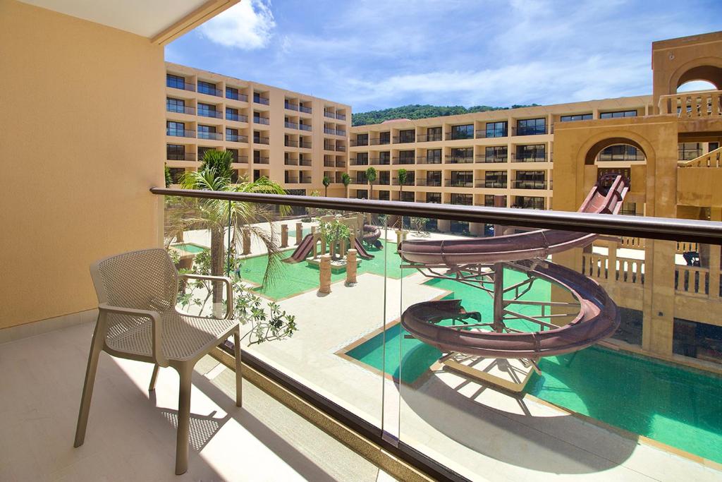 Balkons ar skatu uz viesnīcas baseinu un ūdens slidkalniņiem, apzaļumots pagalms un saulains laiks.