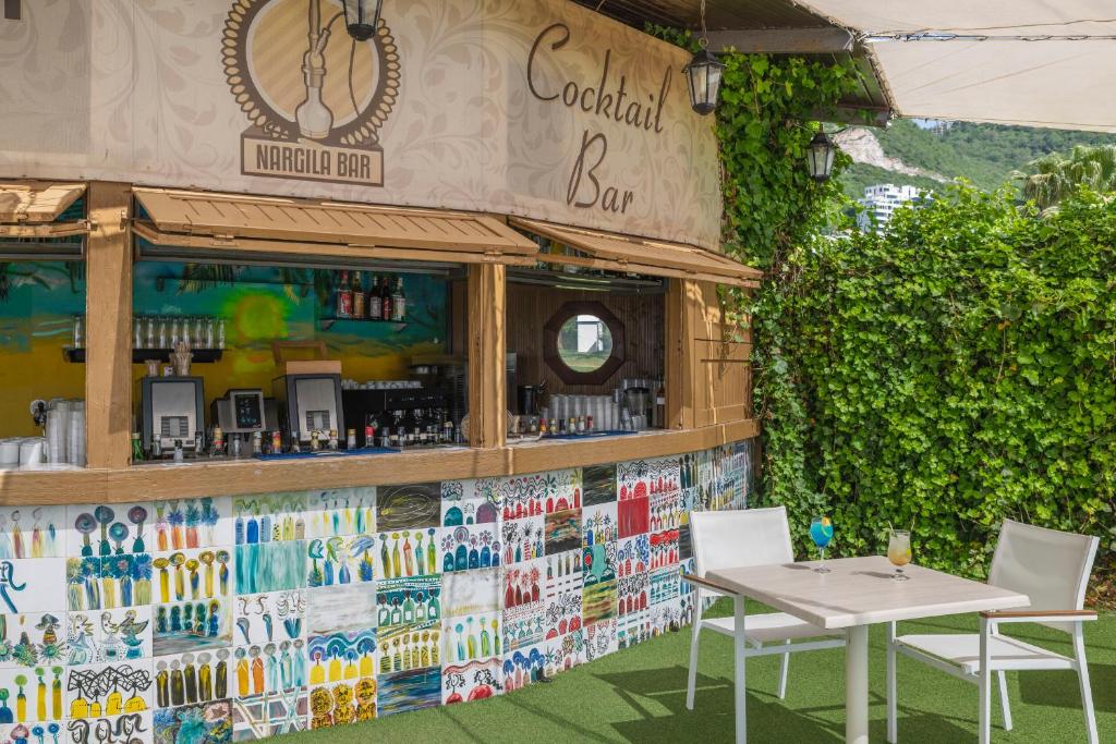 Nargila bārs ar krāsainu flīžu sienu un kokteiļu galdiem, kas izveidots zaļojošā vidē. Bāra logs ir atvērts, piedāvājot dažādus dzērienus un atmosfēru, kas aicina atpūsties.