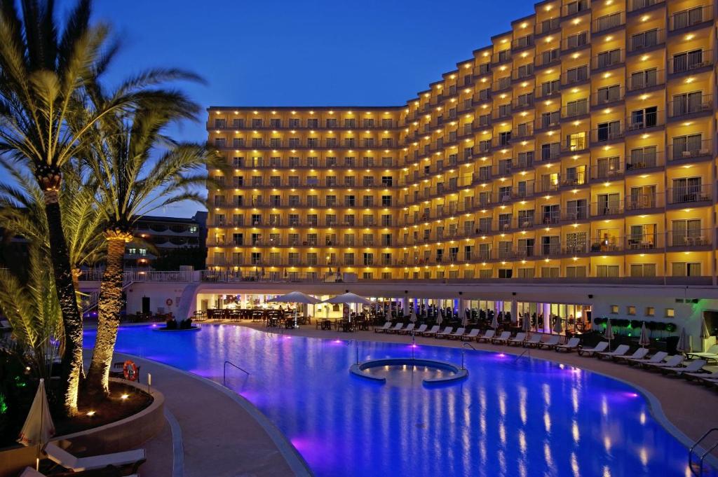 Elegants viesnīcas komplekss ar apgaismotām terasēm un baseinu, kas ieskauj palmām, radot patīkamu atmosfēru vakara stundā.
