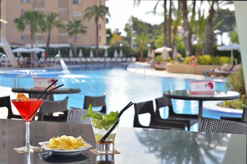 Vasaras atpūtas aina ar divām kokteiļu glāzēm un uzkodu šķīvi, redzot baseinu un palmas fona.