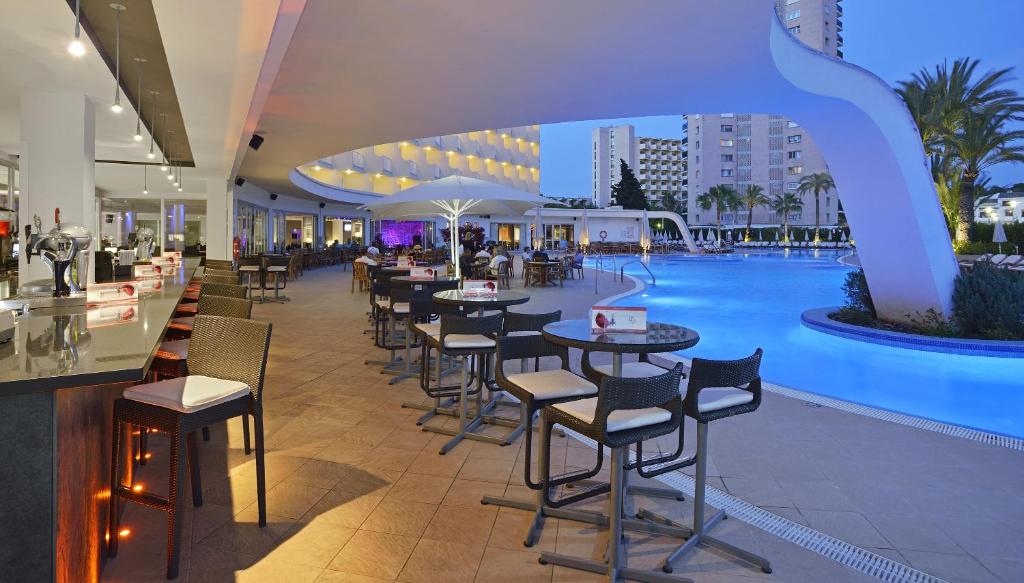 Mūsdienīgs restorāns ar baseinu, kurā ir stilīgas mēbeles un apgaismojums, radot relaksējošu atmosfēru. Apkārt redzamas palmas un viesnīcas ēkas, kas papildina ainavu.