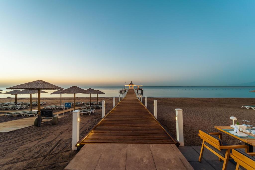 Koka piestātne ved uz mierīgu pludmali, kurā ir sauļošanās krēsli un lietussargi, bet fons ir skaidrs debesis un mierīgs ūdens.