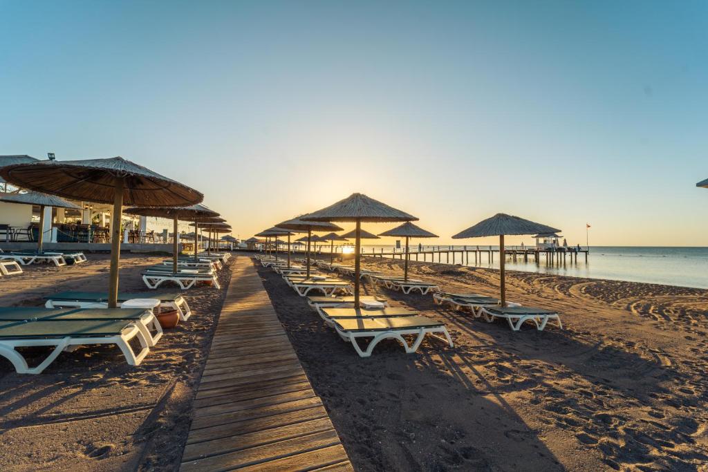 Pludmales skats ar sauļošanās krēsliem un lietussargiem, saulrieta gaismā, kas izgaismo smilšu krastu un piestātni. Ideāla vieta atpūtai un relaksācijai.