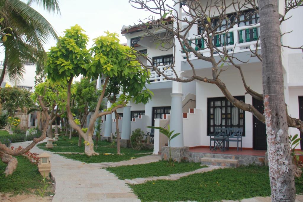 Zaļš dārzs ar koku rindām, kas apņem baltas ēkas ar tumšām logu rāmēm. Ceļš ved cauri dārzam uz ēkām, radot mierīgu atmosfēru.