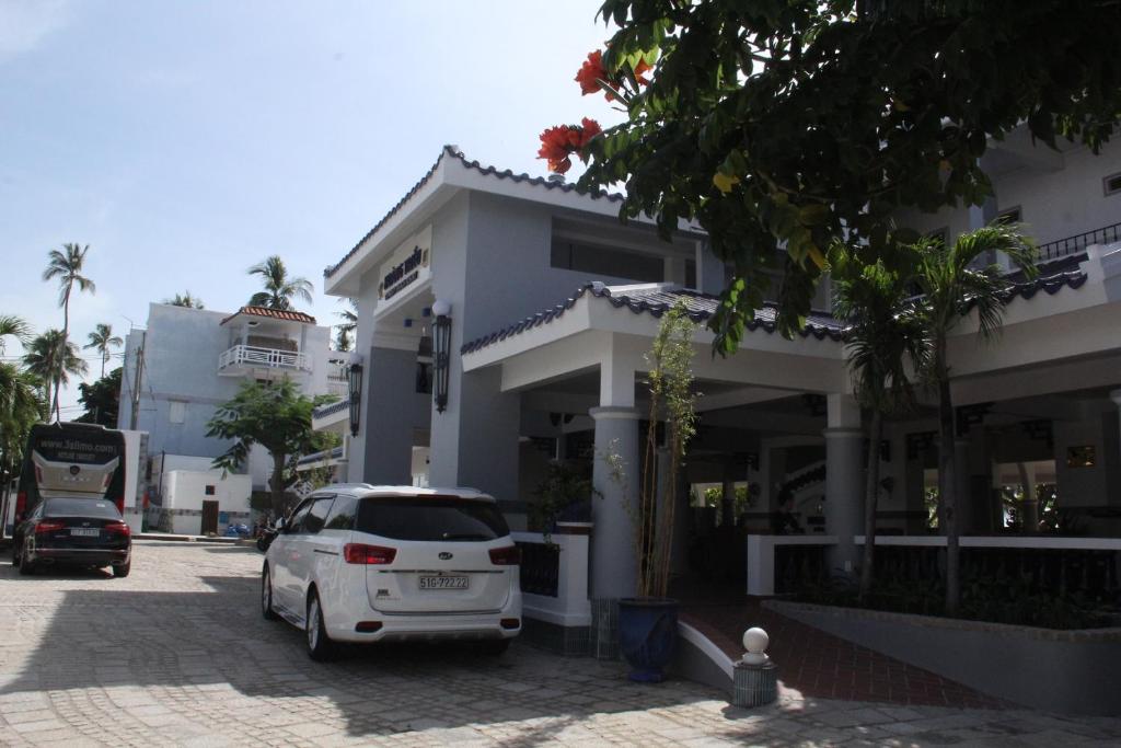 Mūsdienīga viesnīca ar baltiem un pelēkiem arhitektūras elementiem, apzaļumotu teritoriju un automašīnām pie ieejas. Pilsētas ainava ar palmām un saulainu laiku.