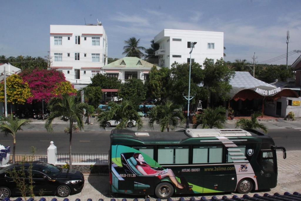 Pilsētas skats ar krāsainām ziedu krūmām un autobusu, kas reklamē transporta pakalpojumu. Fons veido moderni mājas un restorāns 