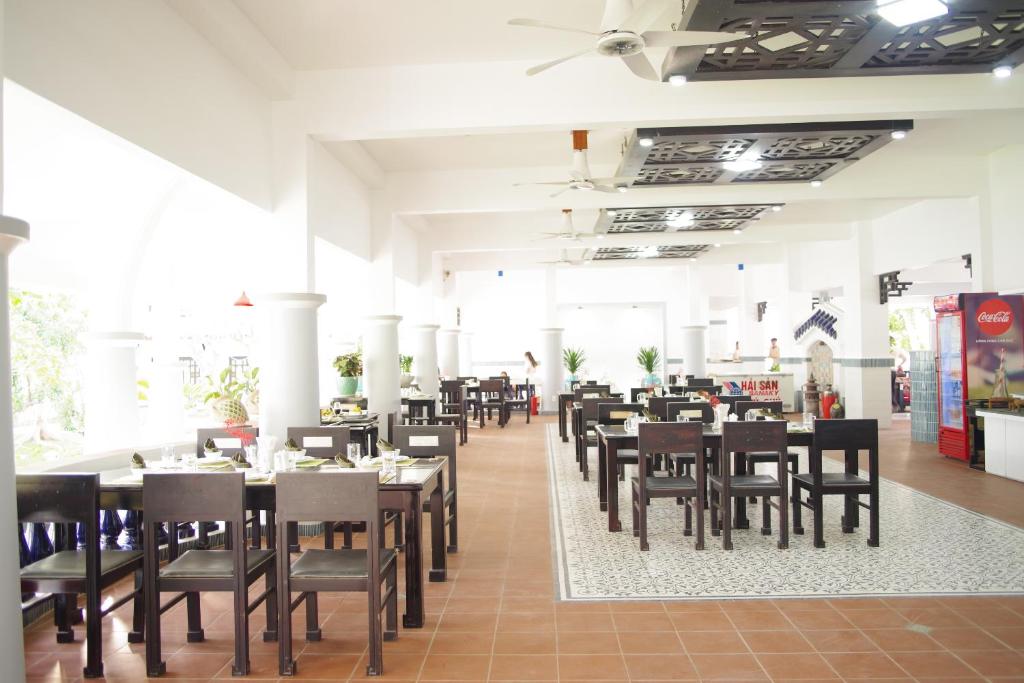 Restorāna interjers ar gaišām sienām un melniem koka galdiem, kur atrodas ēdināšanas zonas un dekoratīvi augi. Telpa ir plaša un gaiša, ar ventilatoriem un tradicionālu grīdas segumu.