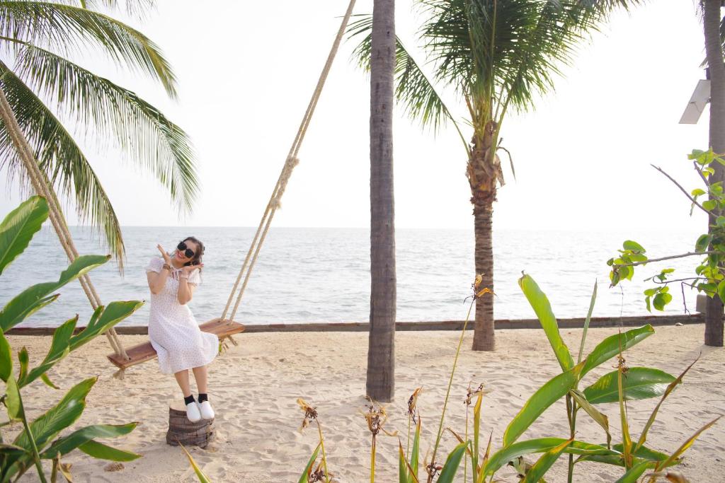 Sieviete baltā kleitā pozē uz šūpolēm pludmalē ar jūru fona. Apkārt palmām un zaļiem augiem.